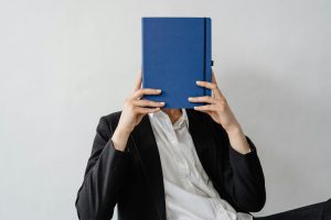Photo of a person sat in a black suit and white shirt with a blue book hiding their face