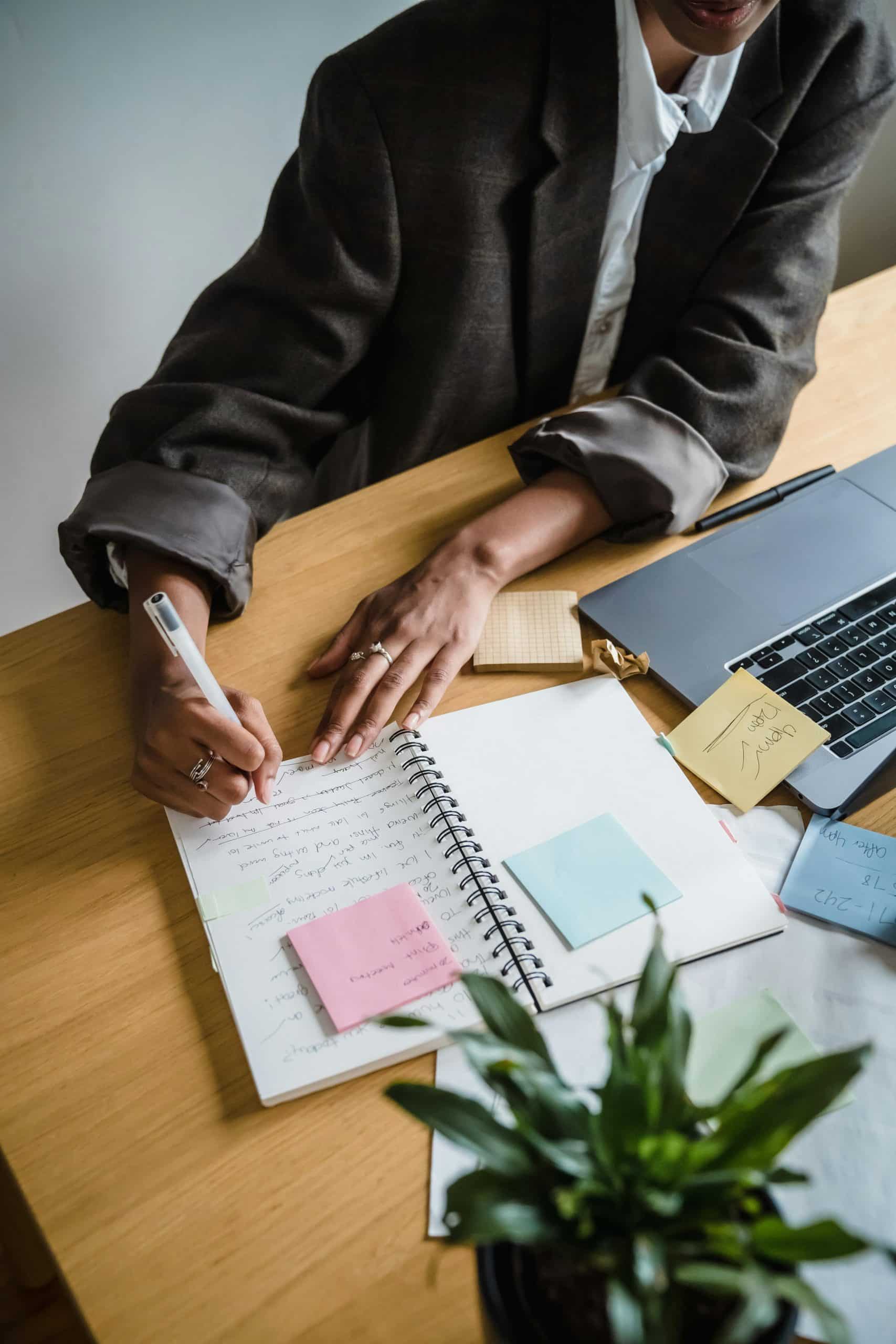 Photo of a woman with her sleeves rolled up writing in an A4 pad that also has post-it notes stuck on. There is an open laptop as she worked through the training provided by The Better Content Club.