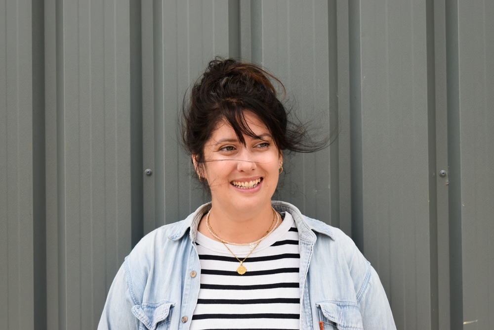 Headshot of Victoria a dark haired woman standing in front of a walk with a striped t-shirt on.